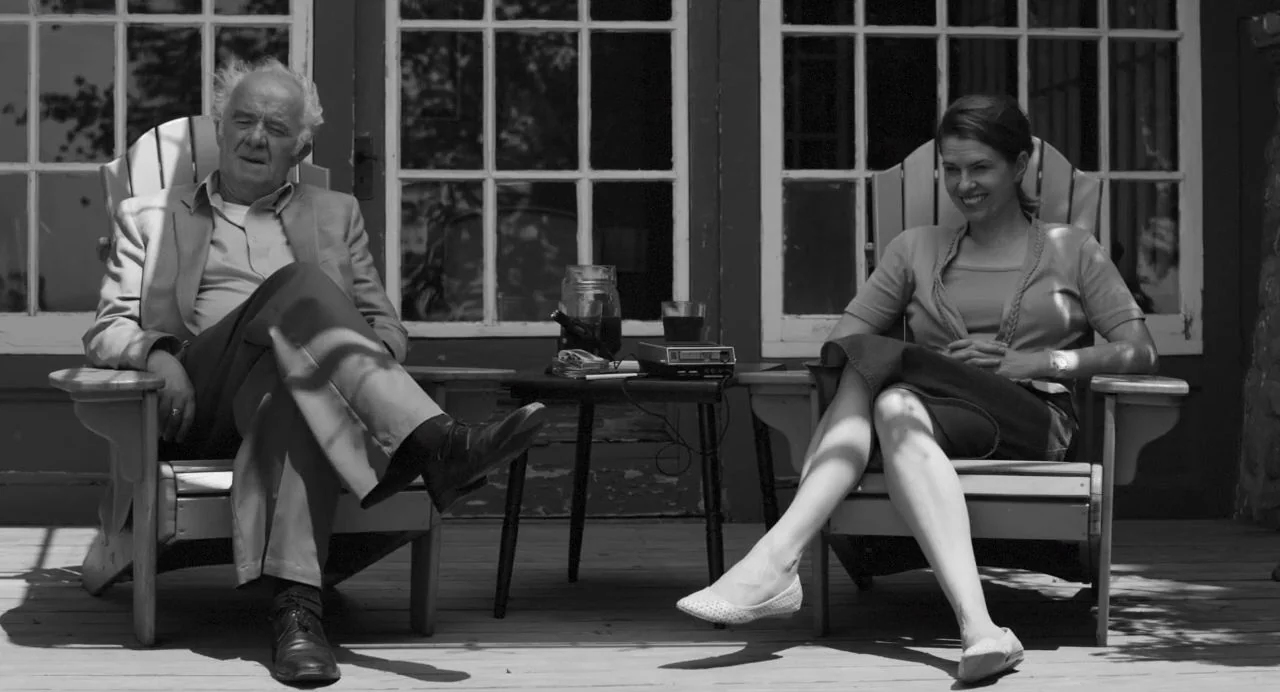 Song of Granite (2017) movie still: fullbody — A wooden deck features two Adirondack chairs positioned in front of a windowed building. An elderly…