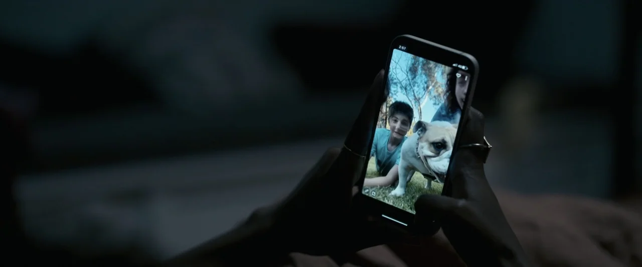 Talk to Me (2023) movie still: closeup — A darkened interior space shows a pair of hands holding a modern smartphone screen. The screen displays a…