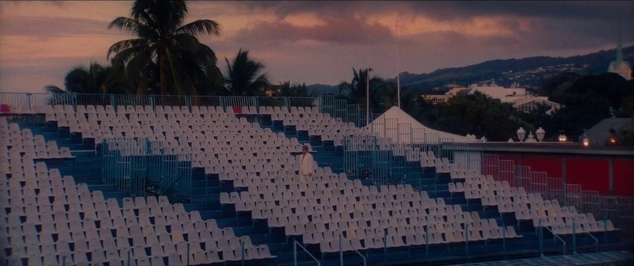 Pacifiction (2022) movie still: wide — A vast outdoor stadium with rows of empty white seats facing an unseen field. A solitary man in a suit stands…