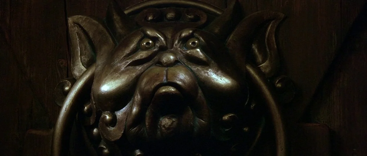 Labyrinth (1986) movie still: closeup — A dark bronze creature-shaped door knocker is mounted on a heavy wooden surface. The sculptural details show…