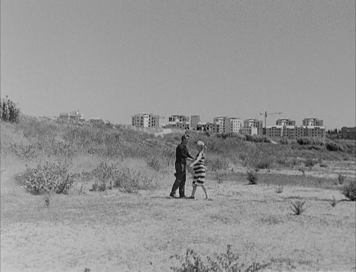 Nights of Cabiria (1957) movie still: wide — An arid, grassy field stretches toward a background of unfinished, multi-story apartment buildings. A…