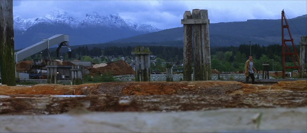 Insomnia (2002) movie still: wide — A sprawling industrial lumber yard features massive log piles and wooden pier pilings. A man walks across the…