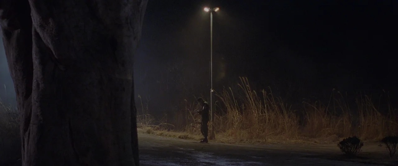 Joint Security Area (2000) movie still: wide — A dark, empty paved area is illuminated by a single tall streetlamp at night. A person stands beneath…