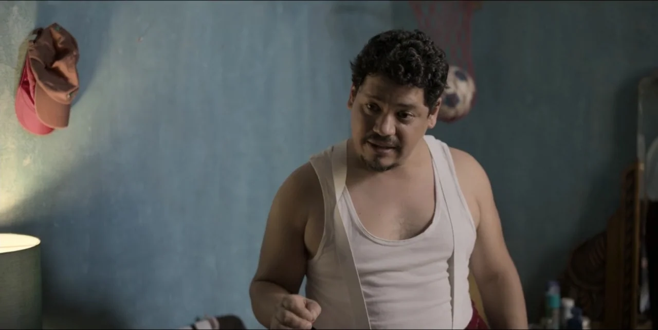 El sabor de la Navidad (2023) movie still: medium — A dimly lit room with blue walls and a small wall-mounted basketball hoop. A man in a white tank…