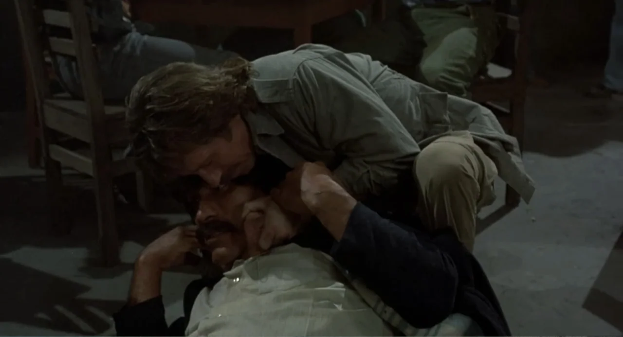 Firewalker (1986) movie still: medium — A dimly lit room contains two men grappling on the floor near wooden furniture. One man kneels over the…