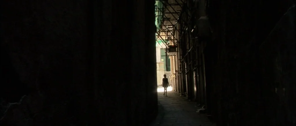 Genova (2008) movie still: wide — A narrow stone alleyway is lined with dark walls and metal scaffolding structures. A small child walks away from…