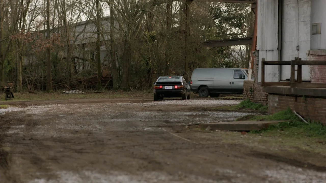 Die Hart (2023) movie still: wide — A muddy dirt lot sits before a gray warehouse building with a loading dock. A black sports car and a silver van…