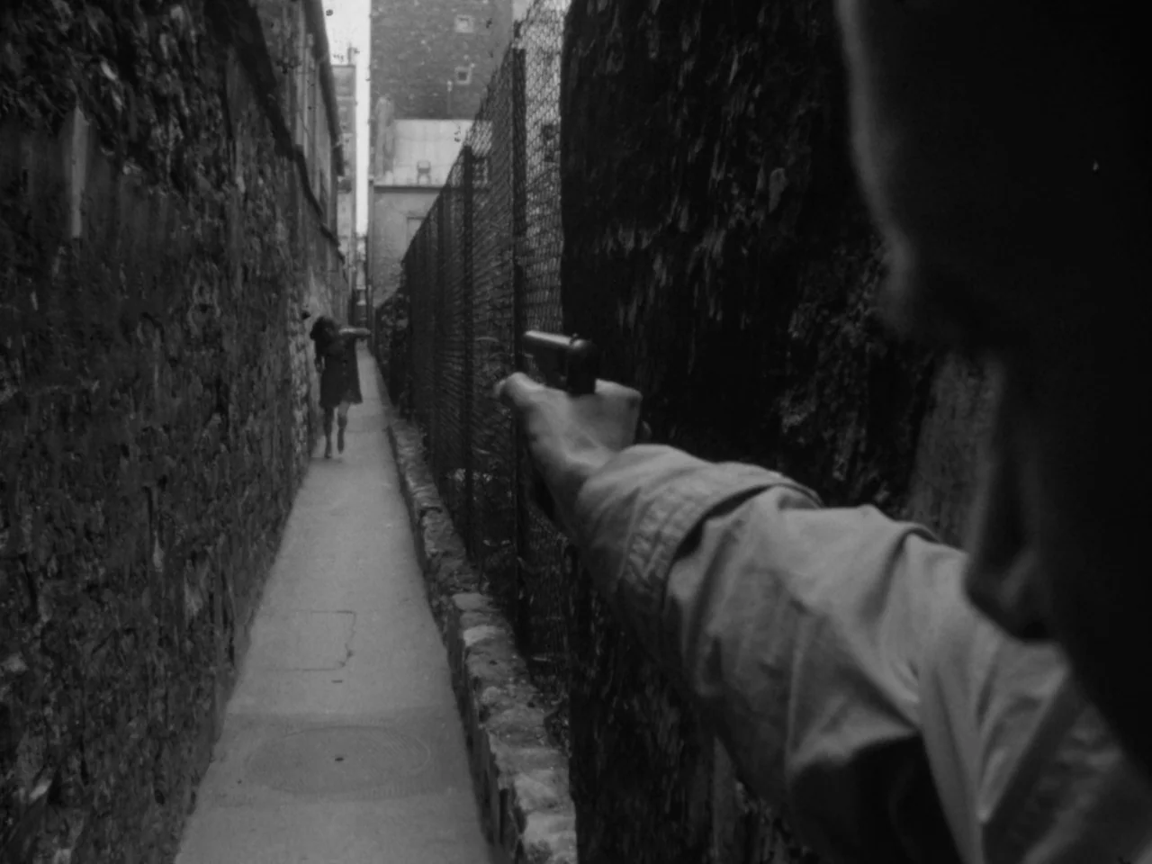 A Woman Kills (1968) movie still: medium — A cramped, concrete-walled outdoor passage is defined by a metal mesh fence. A foreground figure aims a…