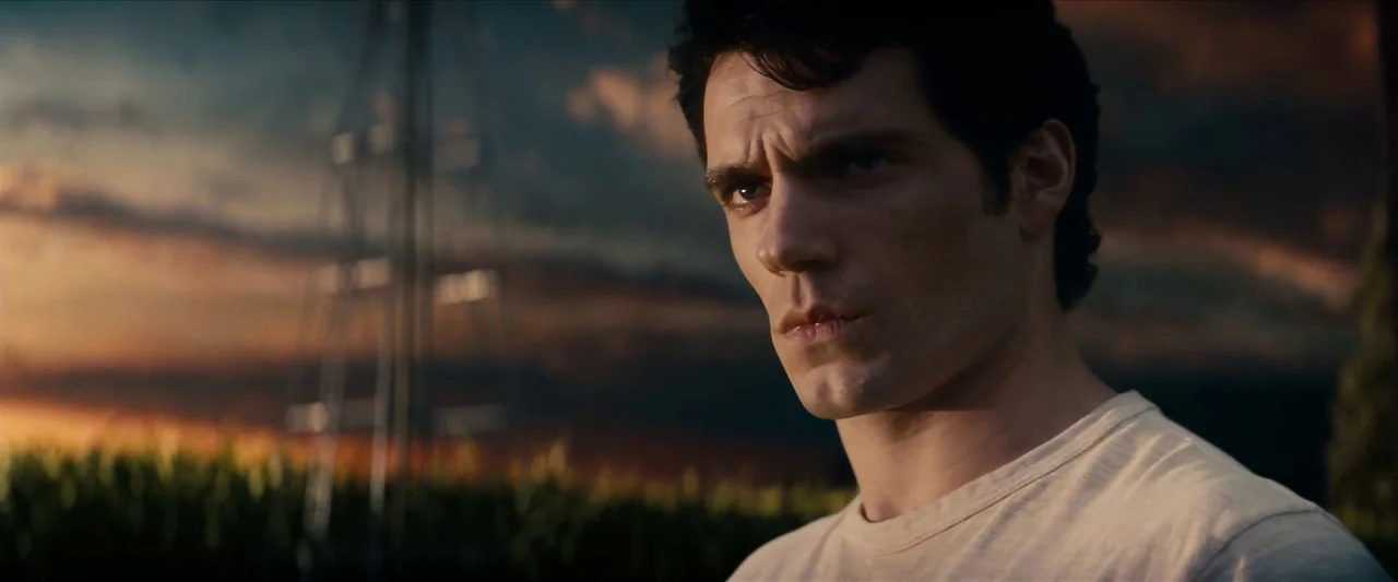 Man Of Steel (2013) movie still: closeup — A man stands in a cornfield under a dim, cloudy twilight sky. He looks past the frame while the sun sets…