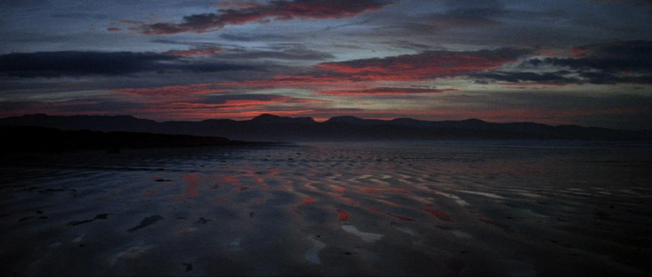 Macbeth (1971) movie still: establishing — A dark, wide beach with rippled wet sand stretches toward a distant mountain silhouette. Red-tinged…