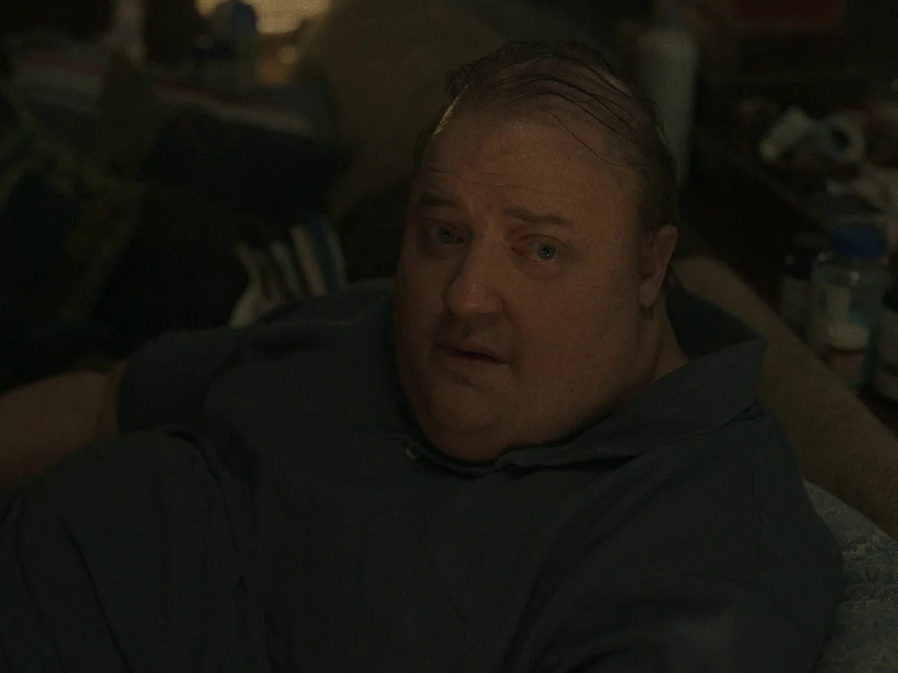 The Whale (2022) movie still: closeup — A dimly lit room containing cluttered furniture and various bottles in the blurred background. An obese…