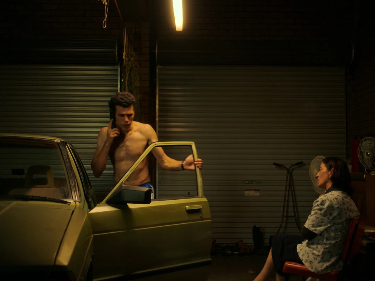 Of An Age (2022) movie still: medium — A dark garage features a pale green vehicle with an open driver's door. A shirtless man uses a mobile phone…