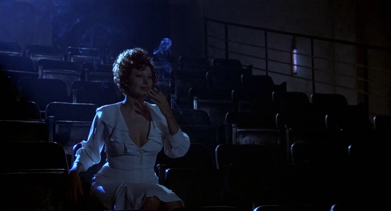 Amarcord (1973) movie still: medium — A dark, empty movie auditorium filled with rows of black folding seats. A woman in a white dress sits in a…