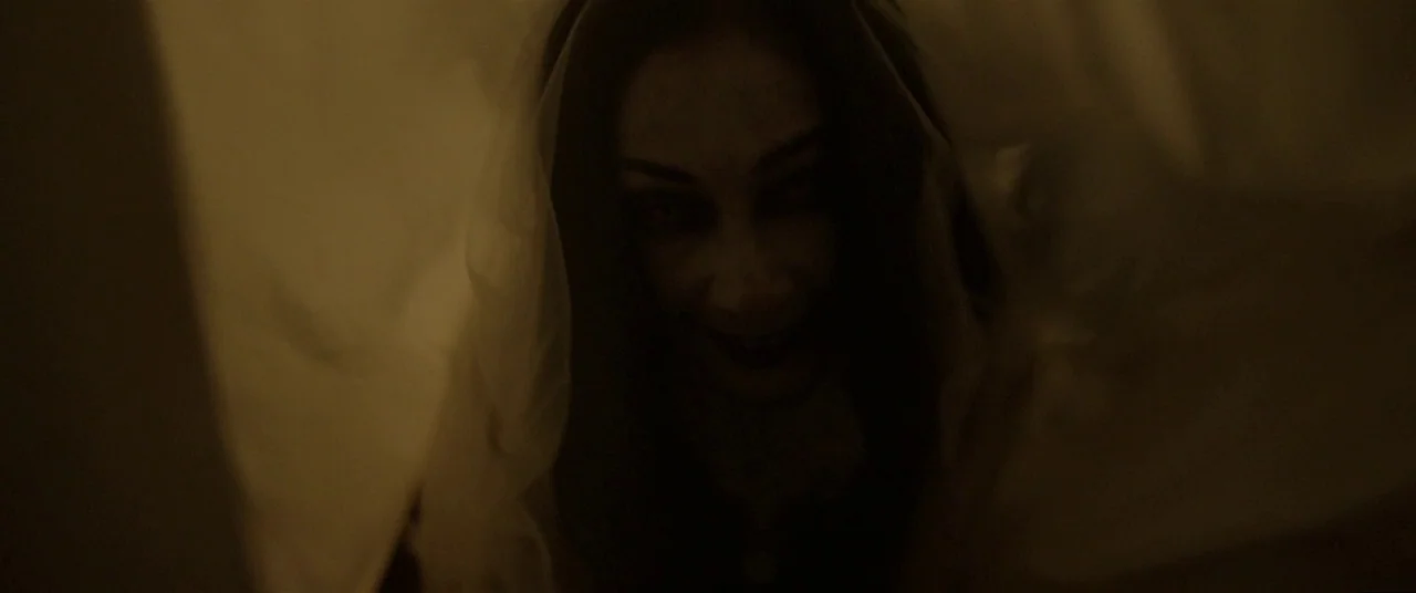 Satan’s Slaves (2017) movie still: closeup — A woman stares from beneath a white sheet in a dimly lit space. She is positioned partially obscured by…
