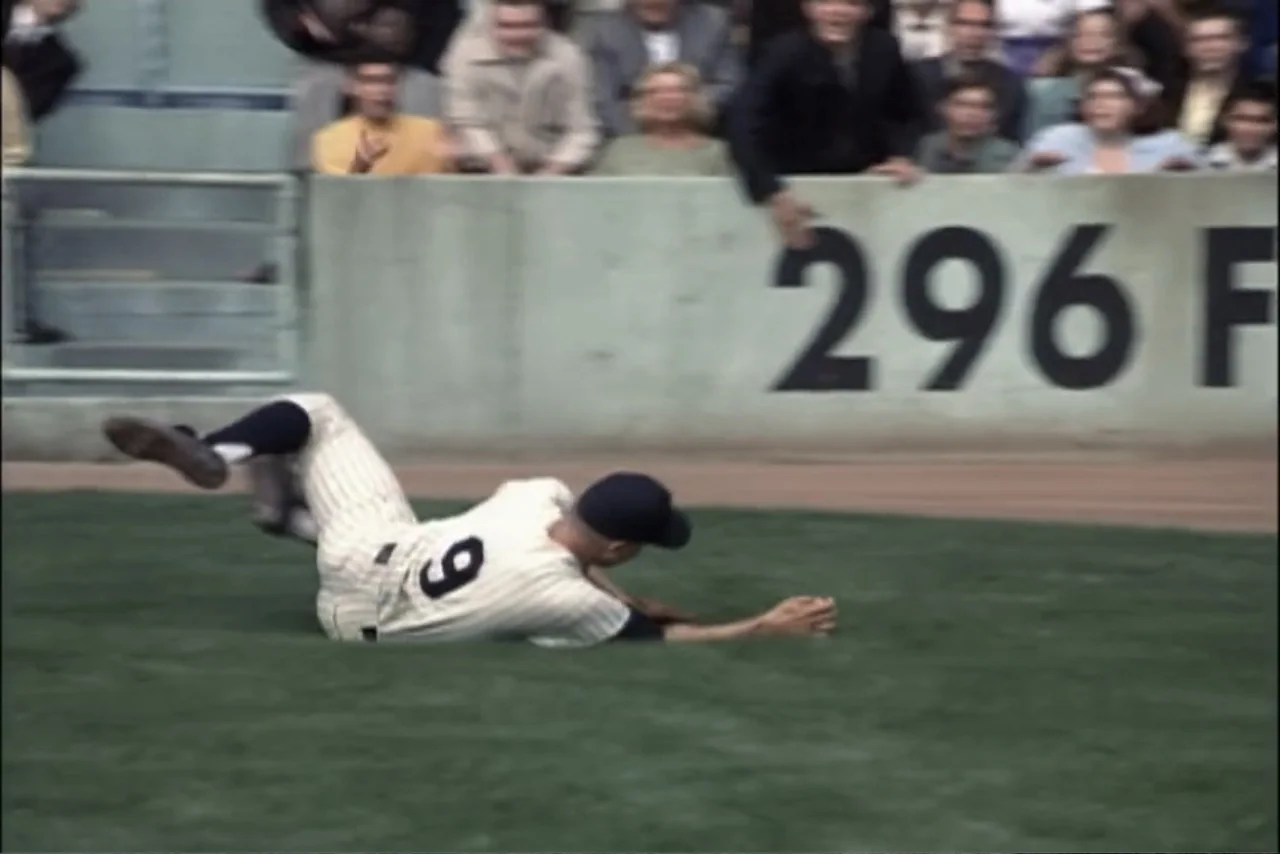 Unification of Japan 61 (2024) movie still: medium — A baseball player slides on green turf before a low concrete outfield wall. The player wears a…