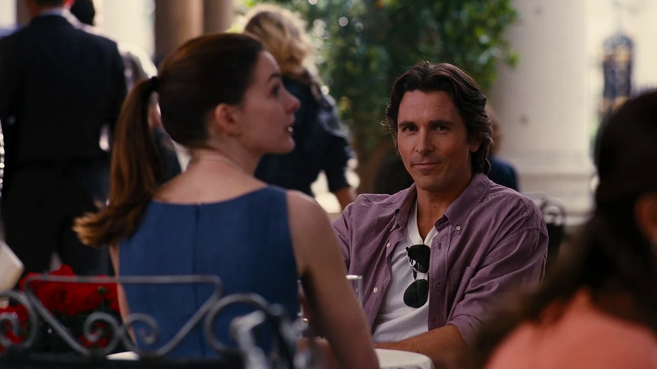 The Dark Knight Rises (2012) movie still: medium — An outdoor cafe terrace with blurred patrons and pillars in the background. A man and woman sit…