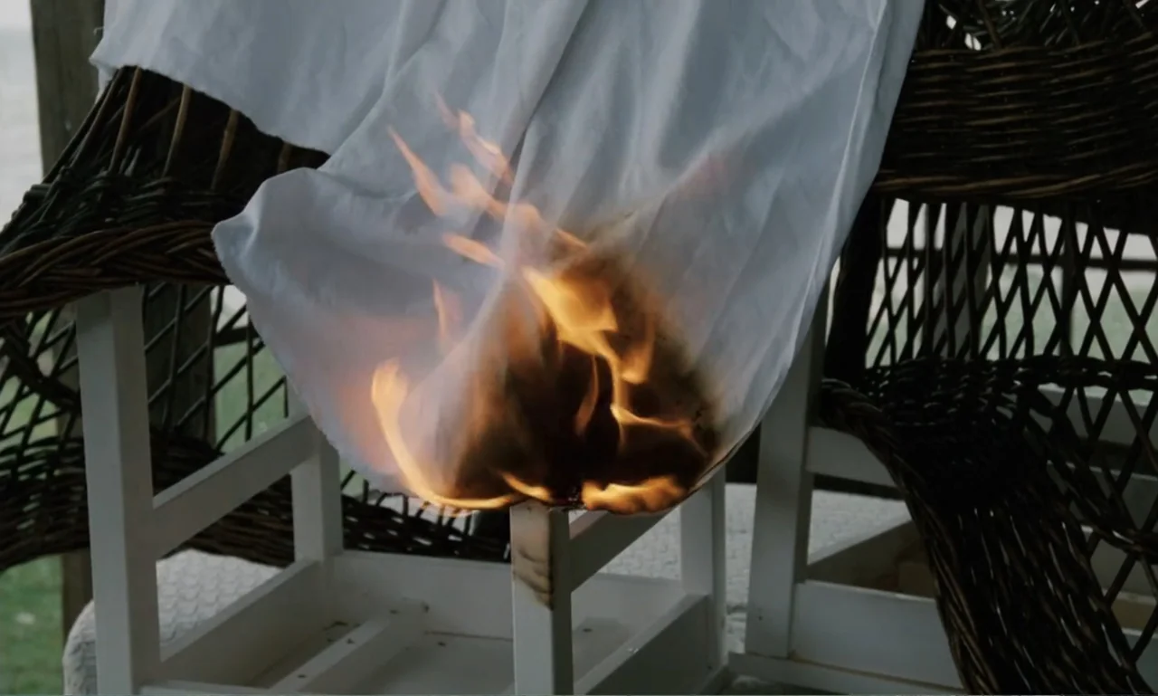 The Sacrifice (2015) movie still: closeup — A wooden chair sits on a porch with a white cloth burning in the center. The fabric is actively catching…