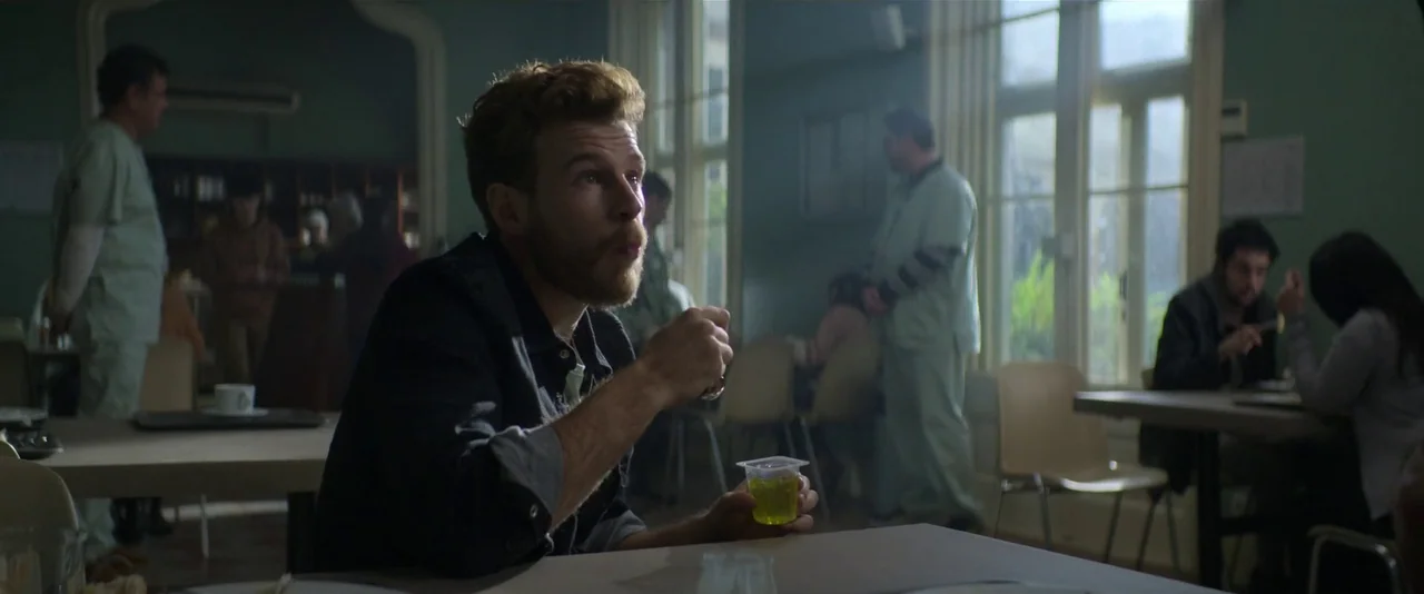 Crazy About Her (2021) movie still: medium — A quiet institutional cafeteria has pale green walls and several tables with occupants. A bearded man…