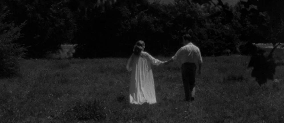 Les Amants (1958) movie still: fullbody — A grassy field with a thick row of dark trees in the distance. A man and a woman in white clothes walk…