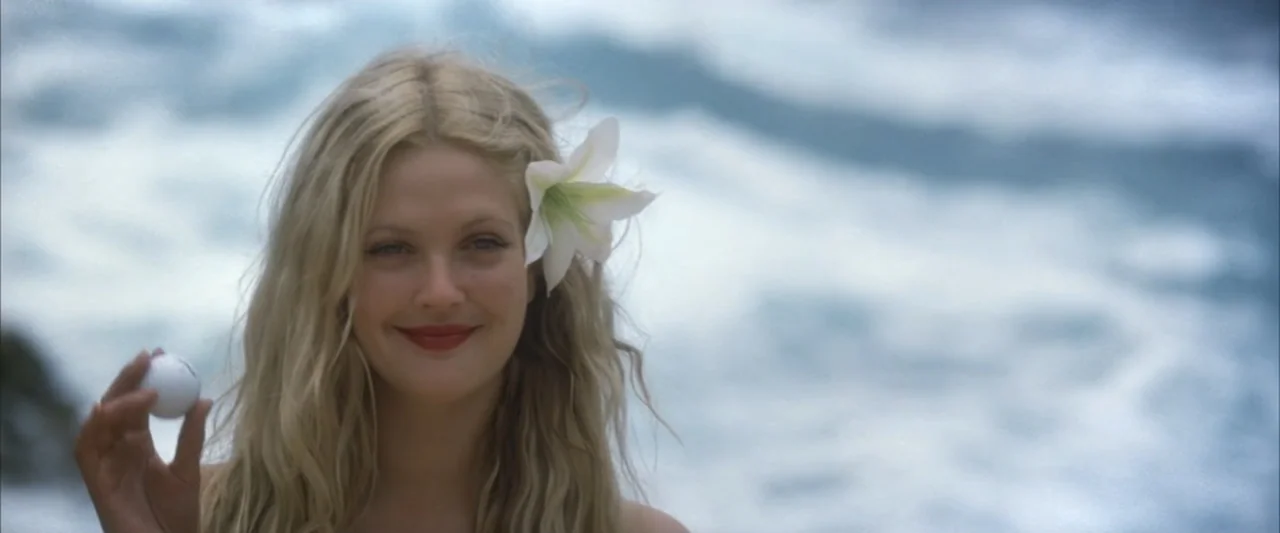 50 First Dates (2004) movie still: closeup — A woman stands before a blurred ocean with crashing waves under bright daylight. She holds a white golf…