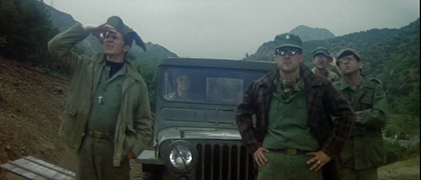 MASH (1970) movie still: wide — A group of soldiers stands by a military jeep on a dirt mountain road. They look up at the overcast sky as if…