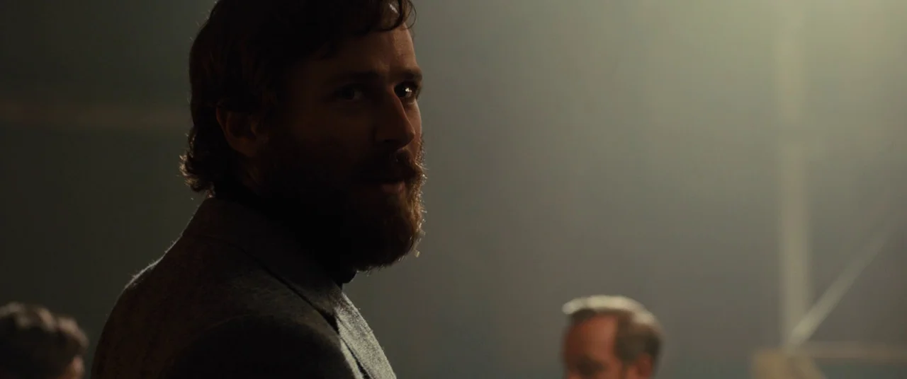 Free Fire (2016) movie still: medium — A dimly lit, hazy interior space with indistinct architectural details in the background. A bearded man in a…