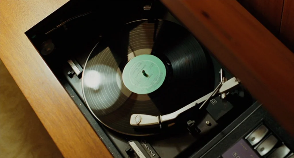A Serious Man (2009) movie still: closeup — A vintage wood-encased record player sits open with a vinyl spinning on the platter. The metal tonearm…