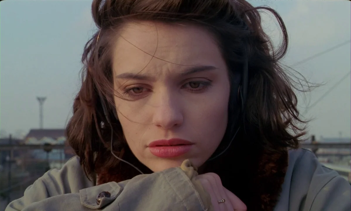 Betty Blue (1986) movie still: closeup — A woman stands outdoors on a bridge during the day. She wears headphones and rests her chin on her hands.
