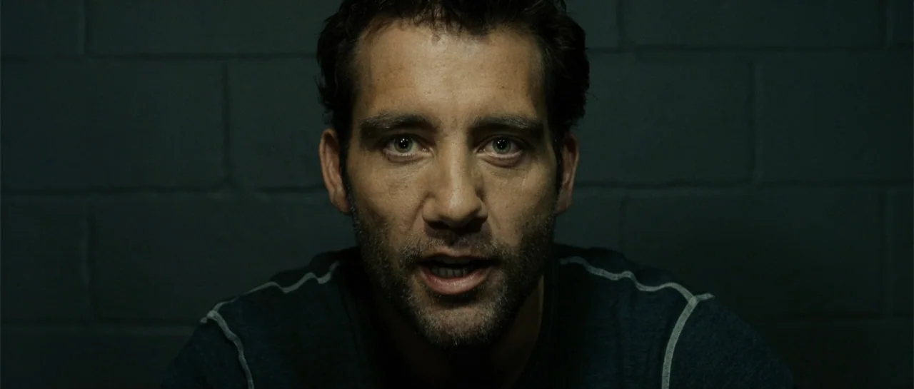Inside Man (2006) movie still: closeup — A dimly lit room features a textured dark grey cinder block wall background. A man faces the camera…
