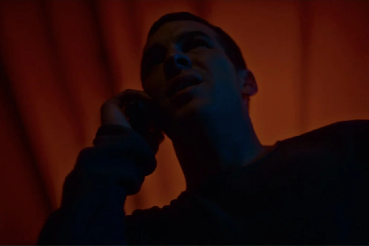 Cross the Line (2020) movie still: closeup — A dimly lit interior space features a backdrop of solid, warm orange fabric. A man holds a dark…