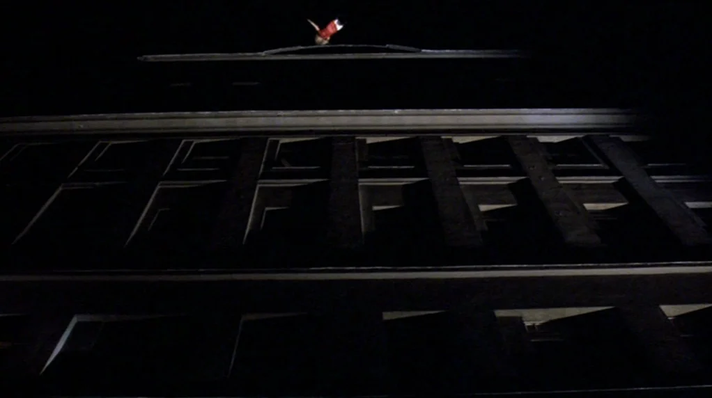 Malina (1991) movie still: wide — A dark, multi-story building exterior at night towers over the camera. A single figure is visible standing on the…