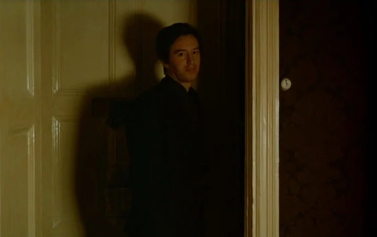 Giliap (1975) movie still: medium — A man in a dark suit stands in a doorway between two rooms. The space features white paneled doors and dark…
