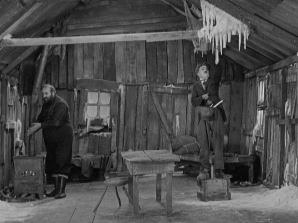 The Gold Rush (1925) movie still: wide — A rustic log cabin features rough wooden plank walls and a slanted ceiling with icicles. One man stands on…
