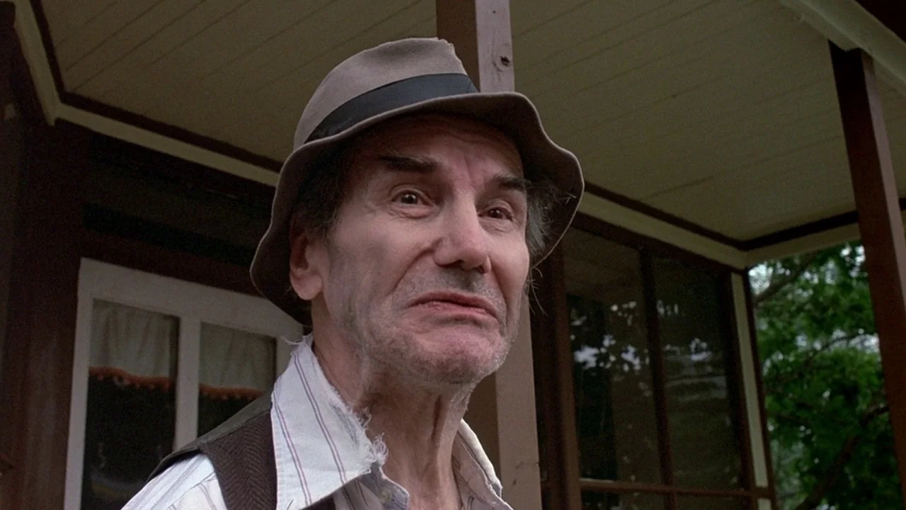 Friday the 13th (1980) movie still: closeup — The porch of a residential house features painted wooden posts and white trim. An elderly man in a…