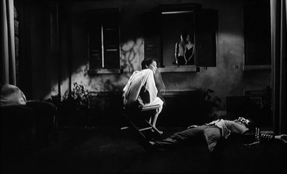 Boy Meets Girl (1984) movie still: wide — A dark night scene at a stone courtyard featuring a building with open windows. A woman sits on a stool…
