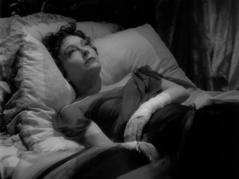 Sunset Boulevard (1950) movie still: medium — A woman lies on a bed with ruffled white pillows and lace bedding. She is resting on her back with…
