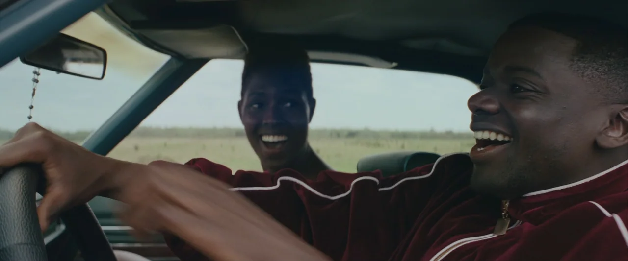 Queen & Slim (2019) movie still: medium — Two people sit inside a moving car driving through a grassy field. They are laughing together while…