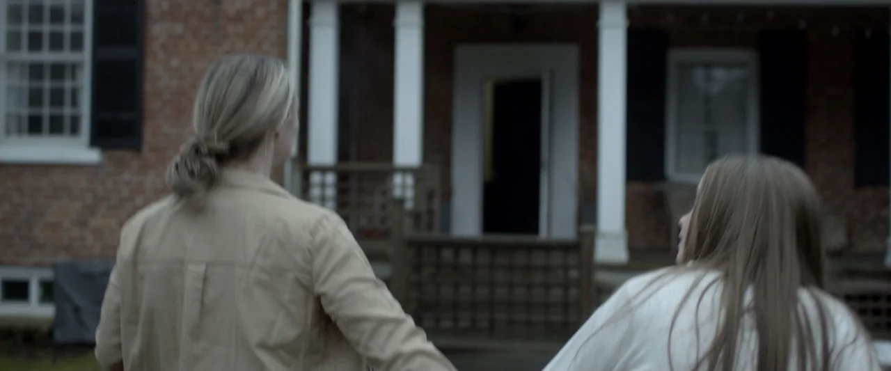 Motherly (2022) movie still: medium — A brick residential building with a white porch and dark shutters stands in daylight. Two females stand with…