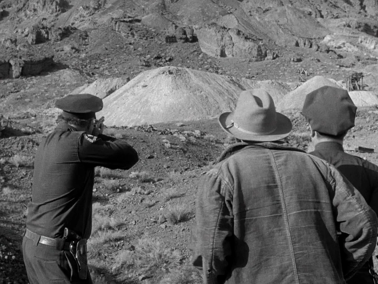 The Prowler (1951) movie still: medium — A rugged desert landscape features rocky mounds and sparse vegetation under direct sunlight. Three men…