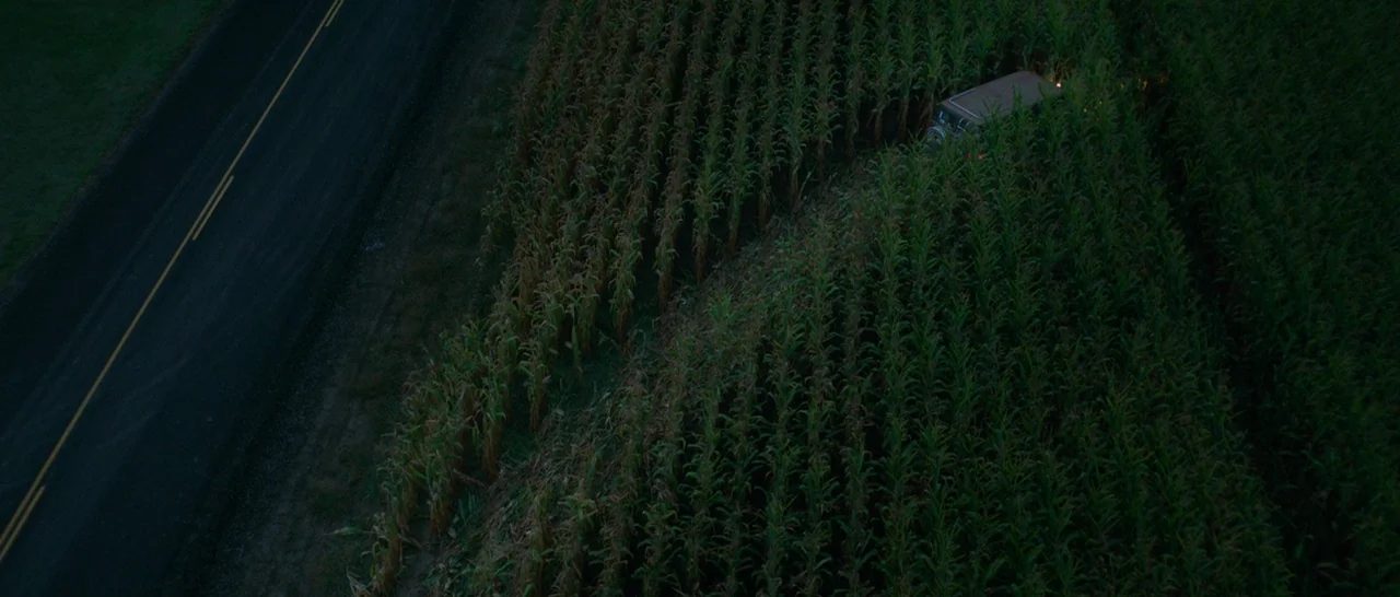 Green Room (2015) movie still: establishing — A dark rural road runs parallel to a dense, overgrown field of tall corn. A dark-colored SUV is…