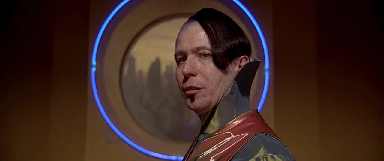 The Fifth Element (1997) movie still: medium — A futuristic room features a circular window displaying a distant landscape under a ring of neon. A…