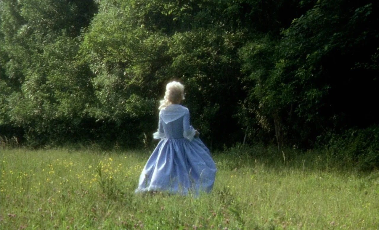 The Beast (1975) movie still: fullbody — A lush grassy meadow borders a dense wall of tall green trees. A woman in a historical blue dress walks…