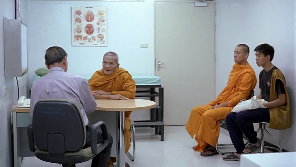 Syndromes and a Century (2006) movie still: wide — A sterile medical office has white walls, a desk, and a medical chart poster. One man in a dress…