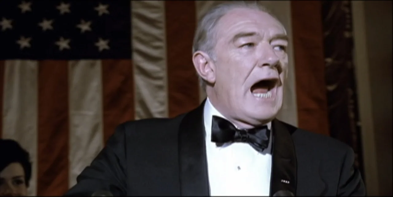 Path to War (2003) movie still: medium — A man stands before a large American flag hanging on a wall indoors. The man in a formal tuxedo speaks…