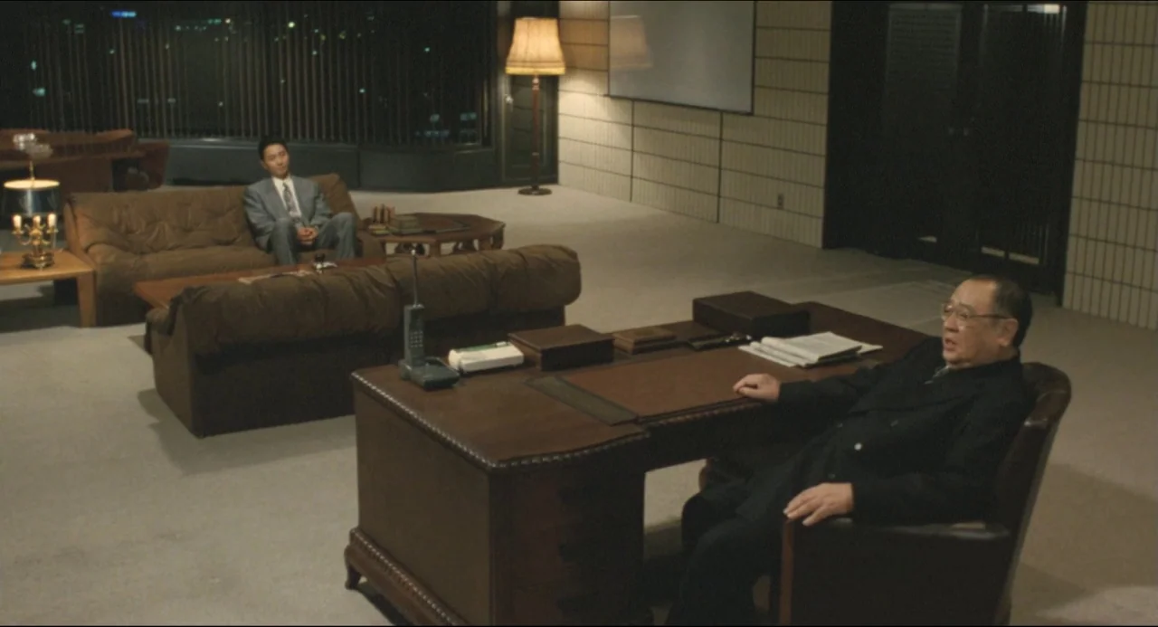 Godzilla vs. Biollante (1989) movie still: wide — A spacious high-floor room has a large mahogany desk and dark leather furniture seating. An older…