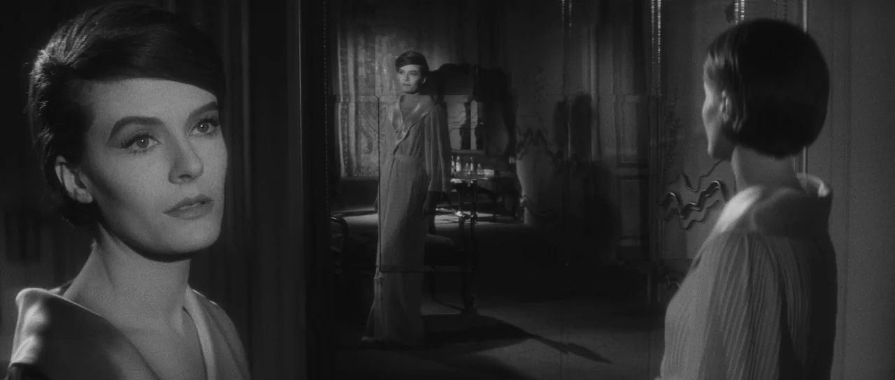 Last Year at Marienbad (1961) movie still: medium — A dimly lit room features a large, ornate mirror reflecting a woman in a robe. The woman stands…