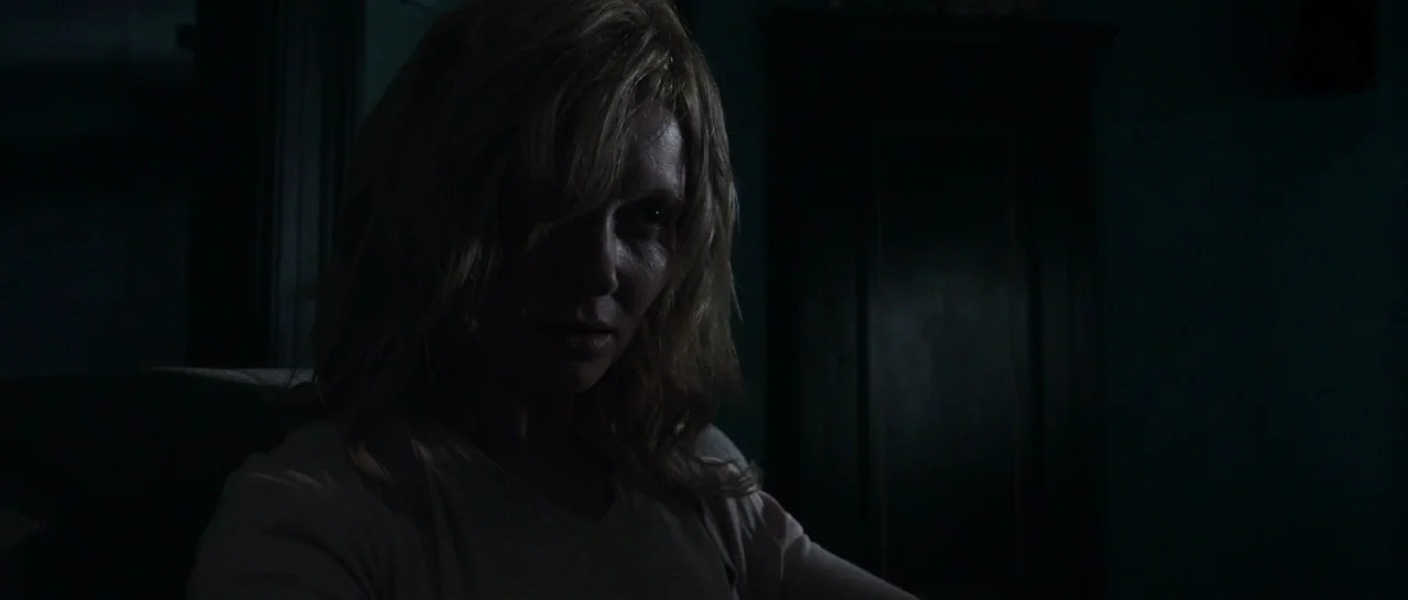 The Babadook (2014) movie still: medium — A dimly lit room provides a dark background for the subject in frame. A woman with blonde hair stares into…