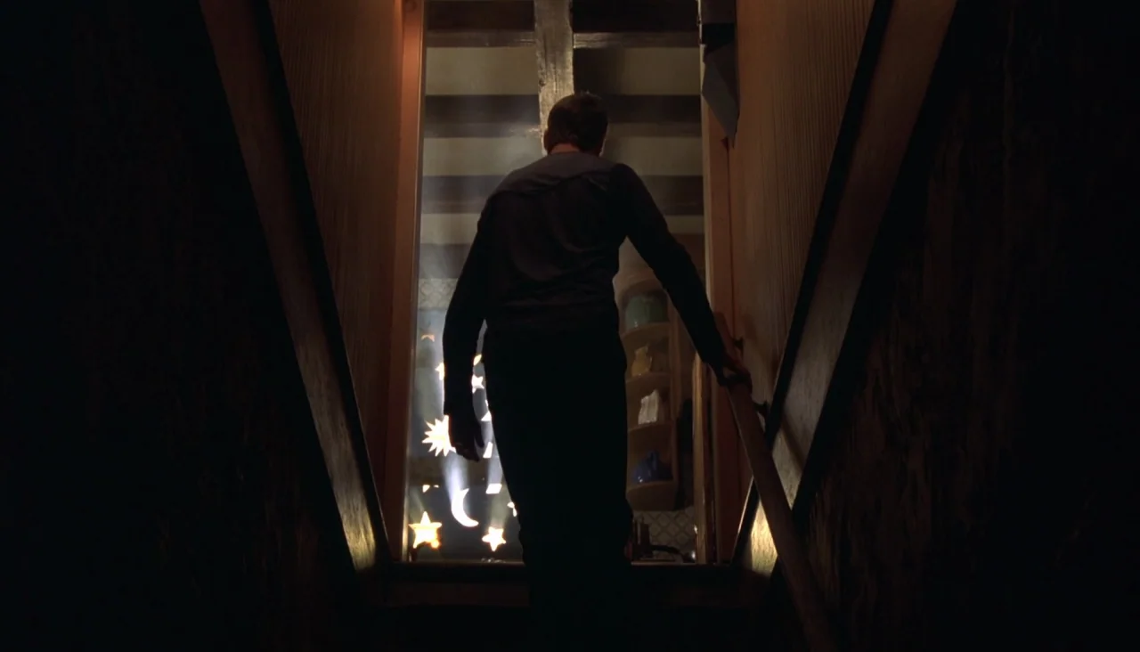 Signs (2002) movie still: fullbody — A dark, enclosed staircase leads upward toward a brightly illuminated opening. The silhouette of a man walks…