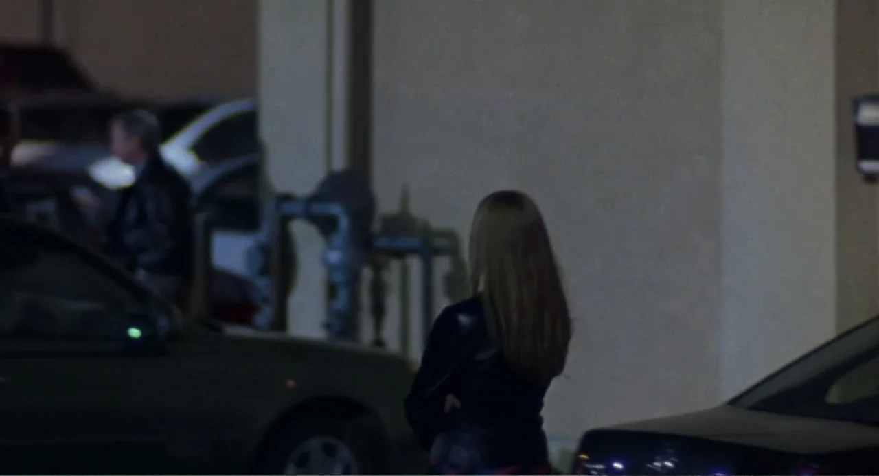 Forty Shades of Blue (2005) movie still: medium — A dimly lit parking lot features a concrete wall and parked vehicles at night. A woman in a…