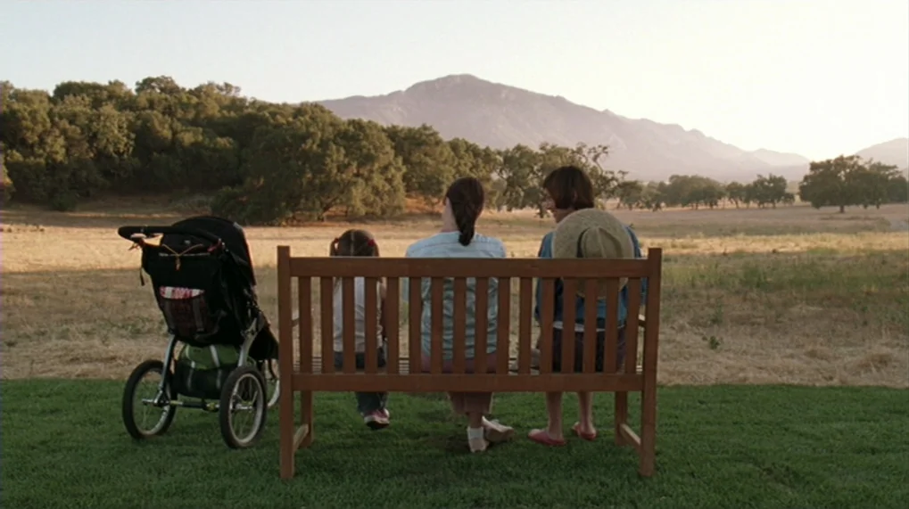 Year Of The Dog (2007) movie still: wide — A wooden bench sits on a grassy lawn facing a wide dry meadow. Three people sit on the bench with a…
